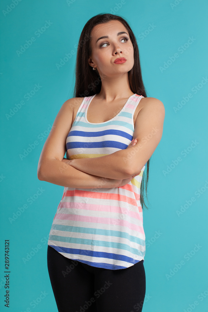 Brunette woman with long hair, dressed in striped shirt and black leggings, posing against blue studio background. Sincere emotions. Close-up. Brunette woman with long hair, dressed in striped shirt and black leggings, posing against blue studio background. Sincere emotions. Close-up.