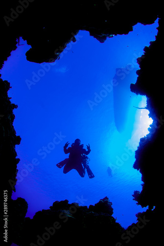 A shot of a scuba diver taken through a window in the coral reef. The silhouette of a dive boat can be seen floating on the surface way above