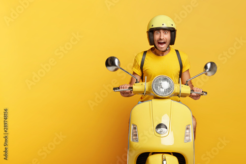 Emotional man drives fast motorbike scooter, reacts emotionally on traffic jam at road, has stupefied expression, carries rucksack on shoulders, wears yellow helmet and t shirt, screams from anger © Wayhome Studio