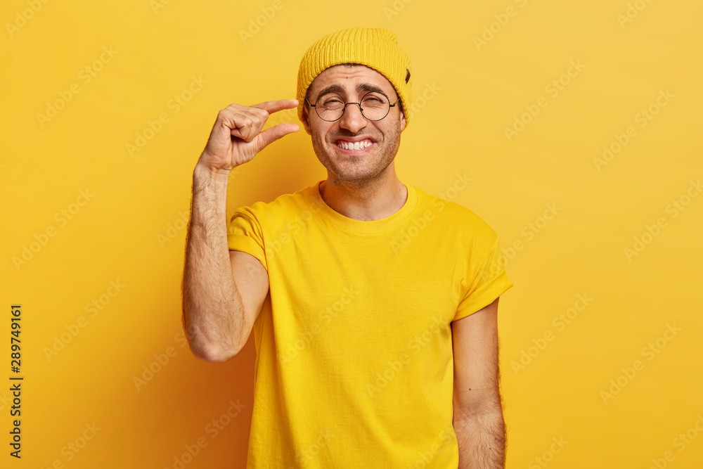 Happy man in casual yellow apparel, makes tiny gesture, shows something very little, smiles gladfully, has cheerful expression, wears transparent spectacles, isolated over yellow wall. People and size Happy man in casual yellow apparel, makes tiny gesture, shows something very little, smiles gladfully, has cheerful expression, wears transparent spectacles, isolated over yellow wall. People and size