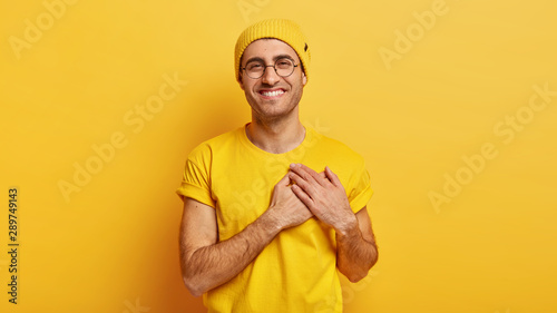 Schilderij op canvas Pleased young man keeps hands on chest near heart, swears about something, makes gratitude gesture, smiles positively, looks at camera with satisfaction, wears casual clothes, models indoor