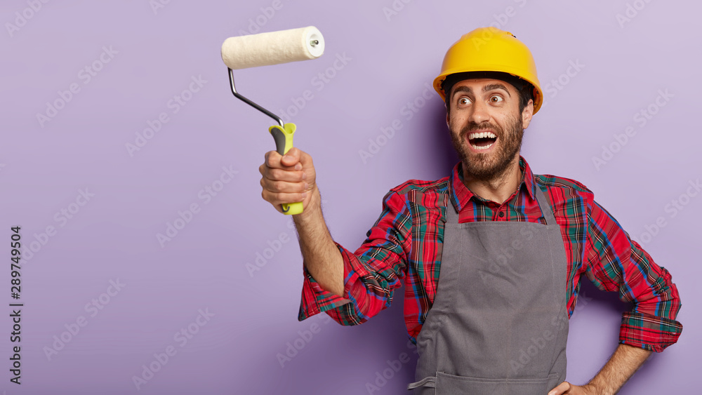 People, repair concept. Happy male builder going to paint walls in house, holds white paint roller, wears special uniform and protective helmet, busy with flat renovation isolated on purple background People, repair concept. Happy male builder going to paint walls in house, holds white paint roller, wears special uniform and protective helmet, busy with flat renovation isolated on purple background
