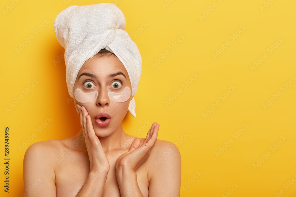 Shocked terrible woman uses new effective cosmetic tool, applies anti wrinkle patches under eyes, stares with stunned expression, wears towel on wet hair, stands topless over yellow background Shocked terrible woman uses new effective cosmetic tool, applies anti wrinkle patches under eyes, stares with stunned expression, wears towel on wet hair, stands topless over yellow background