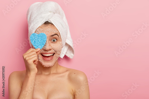 Horizontal shot of happy young female model covers eye with cosmetic sponge, cleans face by natural white sea salt scrub mask, visits spa salon, has sooth healthy skin, naked body, models indoor