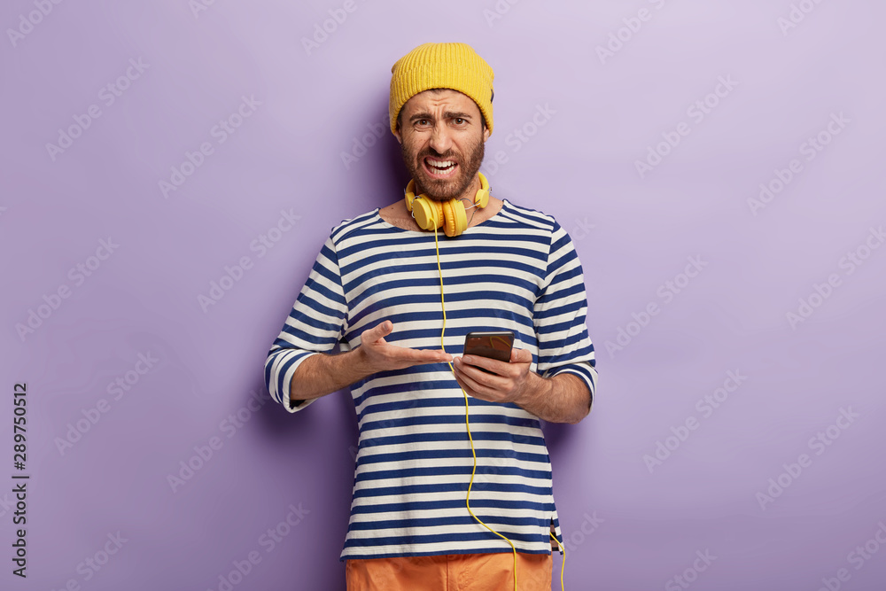Horizontal shot of dissatisfied man points into smartphone device, has unpleasant look at camera, wears stylish clothes, cannot understand how to use new application, isolated on purple background