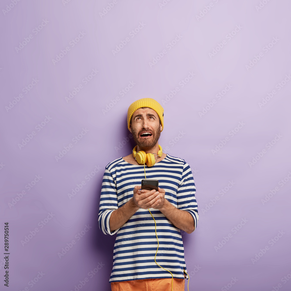 Dissatisfied young man holds modern smartphone, looks above with miserable expression, cannot connect to internet, being always in touch, dressed in fashionable clothes, poses over purple wall Dissatisfied young man holds modern smartphone, looks above with miserable expression, cannot connect to internet, being always in touch, dressed in fashionable clothes, poses over purple wall