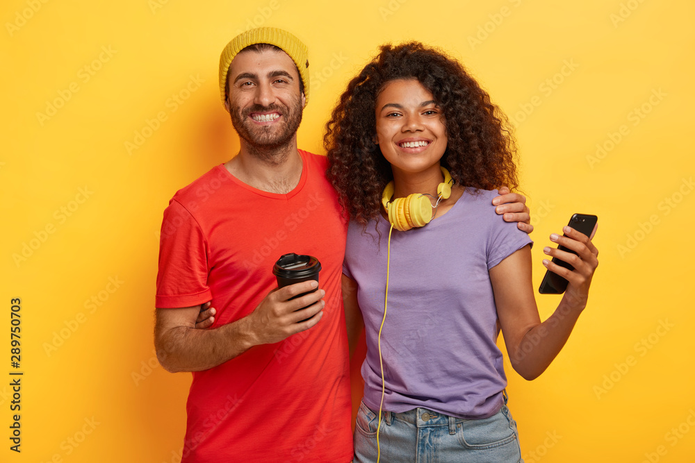 Photo of happy mixed race couple spend free time together, drink coffee and use modern cellular for online communication, dressed in t shirts, stand closely to each other against yellow background Photo of happy mixed race couple spend free time together, drink coffee and use modern cellular for online communication, dressed in t shirts, stand closely to each other against yellow background
