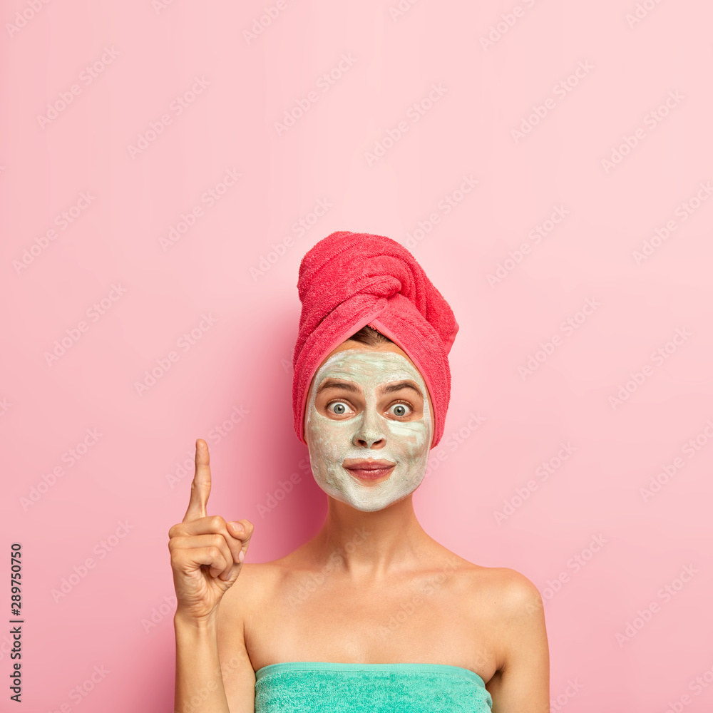 Beauty and spa concept. Photo of lovely woman points index finger above, wears nourising mask, reduces pimples, avoids problems of skin dryness, wrapped in bath towel, isolated on pink wall. Beauty and spa concept. Photo of lovely woman points index finger above, wears nourising mask, reduces pimples, avoids problems of skin dryness, wrapped in bath towel, isolated on pink wall.