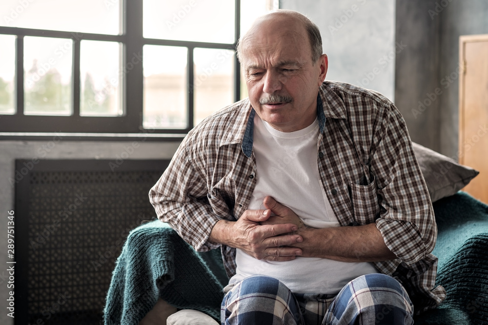Senior hispanic man is having pancreatitis or gastritis or colitis sitting alone at the living room. Poor health at old age concept - obrazy, fototapety, plakaty 