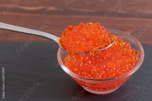 Red caviar in glass jar with silver spoon on the dark brown wooden background