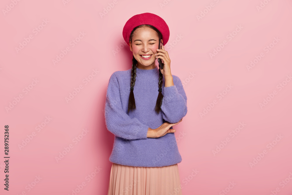 Overjoyed pleased woman with Asian appearance, enjoys funny telephone conversation with friend, keeps modern cellular near ear, has two long pigtails, poses in studio over pink wall. Communication Overjoyed pleased woman with Asian appearance, enjoys funny telephone conversation with friend, keeps modern cellular near ear, has two long pigtails, poses in studio over pink wall. Communication