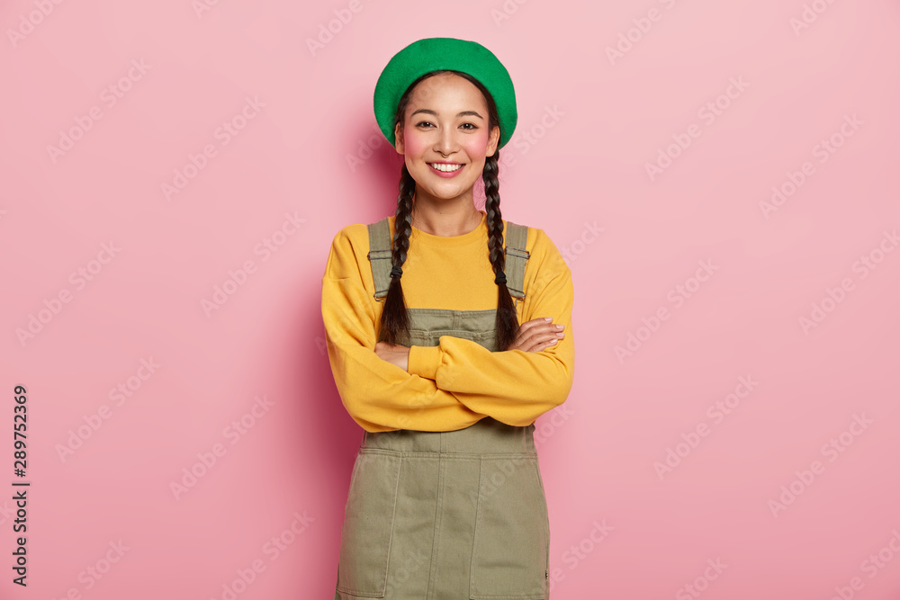 Candid shot of happy Chinese millennial girl keeps arms folded, smiles pleasantly at camera, enjoys pleasant talk with boyfriend, wears green beret, yellow sweatshirt and sarafan, feels joyous Candid shot of happy Chinese millennial girl keeps arms folded, smiles pleasantly at camera, enjoys pleasant talk with boyfriend, wears green beret, yellow sweatshirt and sarafan, feels joyous