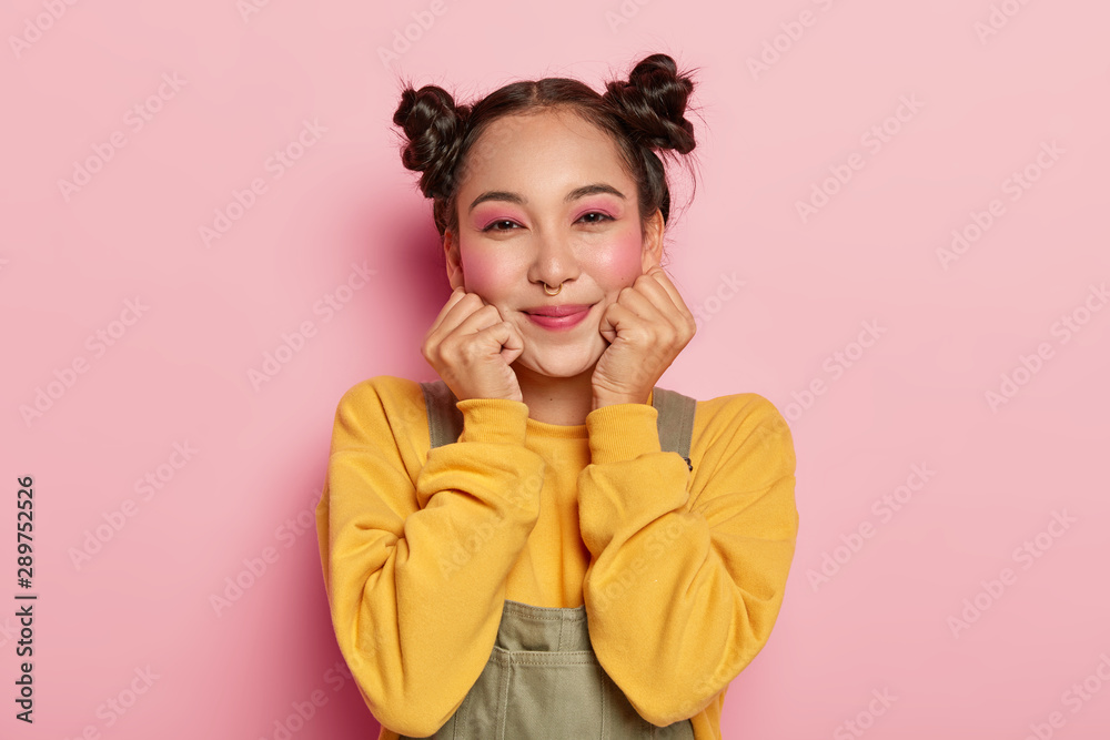 Portrait of happy Asian girl with pinup makeup, dark hair combed in two buns, piercing in nose, wears casual yellow sweatshirt and overalls, stands on pink background, feels happy. Ethnicity, emotions