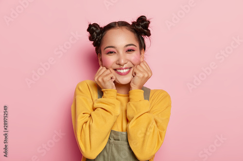 Portrait of beautiful Asian girl with pinup makeup, holds chin with both hands, dressed in casual outfit, has dark hair combed in two buns, poses against rosy background, wears piercing in nose