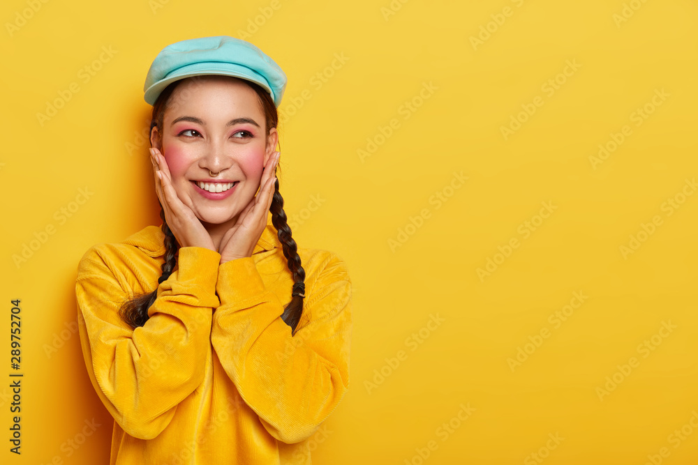 Happy Asian girl touches rouge cheeks, wears makeup and piercing, stylish cap and velvet sweatshirt, has dreamy expression, imagines unforgettable moment with boyfriend, isolated on yellow wall Happy Asian girl touches rouge cheeks, wears makeup and piercing, stylish cap and velvet sweatshirt, has dreamy expression, imagines unforgettable moment with boyfriend, isolated on yellow wall