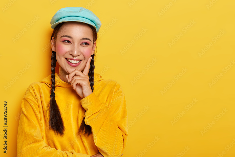 Happy feelings and emotions concept. Beautiful young woman with eastern appearance, touches rosy cheek with index finger, looks aside, has toothy smile, dressed in bright velvet hoody sweater Happy feelings and emotions concept. Beautiful young woman with eastern appearance, touches rosy cheek with index finger, looks aside, has toothy smile, dressed in bright velvet hoody sweater
