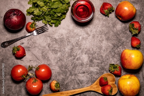 Fresh organic red themed fruit, vegetables, wine, and wooden heart shaped spoon, laying on concrete background countertop. Captured from above. 