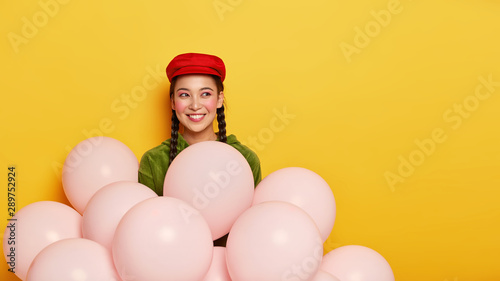 Isolated shot of happy Asian female wears stylish red cap, has dark hair combed in two braids, poses behind airballoons, gets pleasure from awesome party or holiday, looks aside with glad expression
