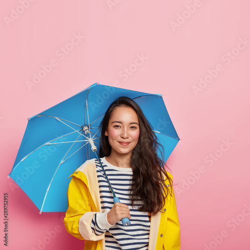 Happy dark haired eastern lady with natural beauty, feels dry and protected, wears waterproof raincoat, carries umbrella, enjoys free time during rainy autumn day. Season and weather concept