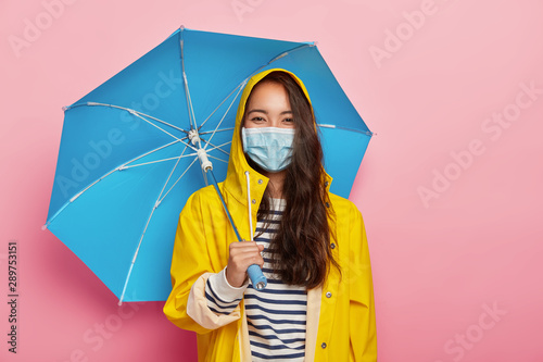 Cuadro en lienzo Photo of Asian woman wears protective mask, faces air pollution during rainy day, stands under umbrella, dressed in yellow raincoat, isolated on pink background