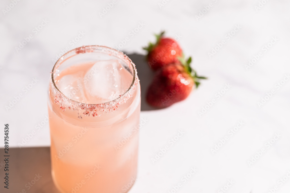 A strawberry lemonade drink in a can like glass standing on a marble countertop. Direct sunlight from the aide casting a hard shadow on the countertop. Bottom is whole strawberry with strawberry sugar A strawberry lemonade drink in a can like glass standing on a marble countertop. Direct sunlight from the aide casting a hard shadow on the countertop. Bottom is whole strawberry with strawberry sugar