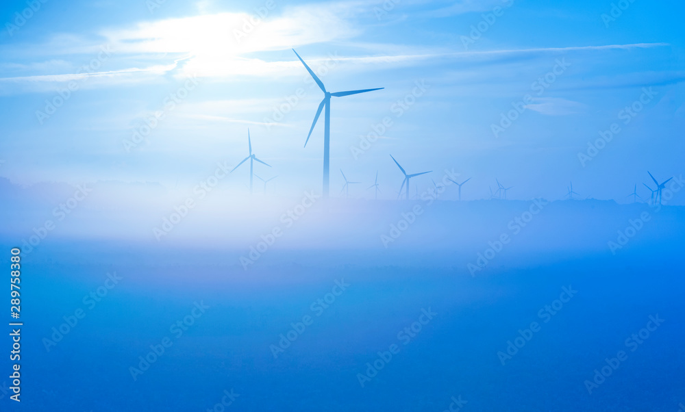 Sun shines on wind turbines in a foggy field at sunrise in summer