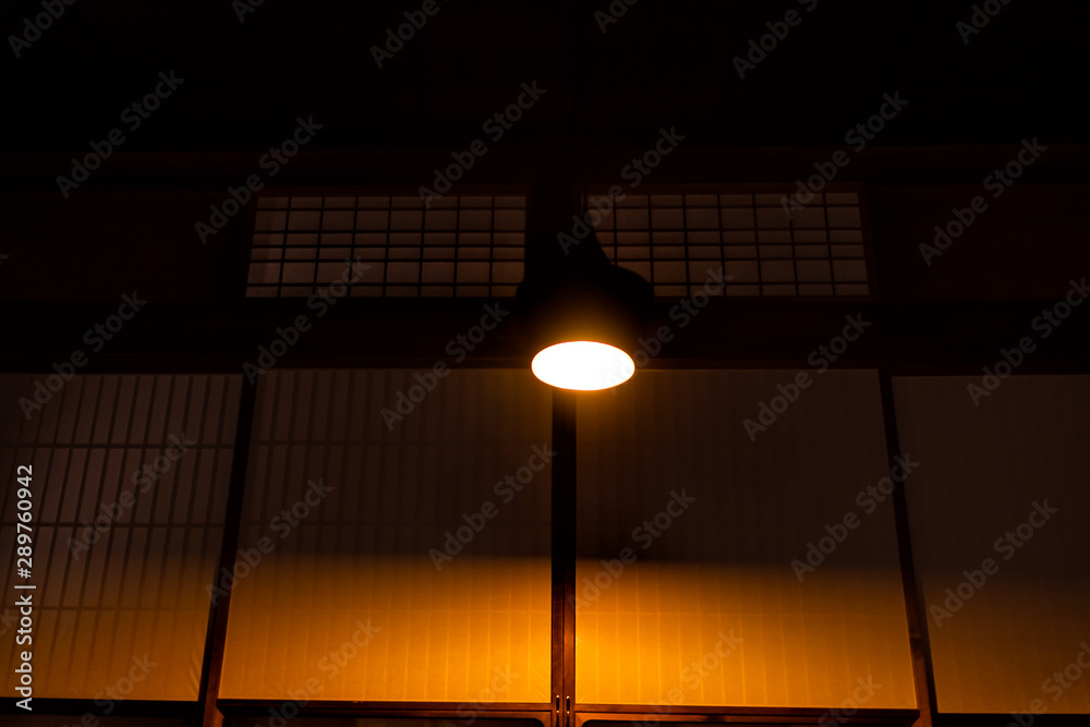 Fototapeta premium Low angle looking up view of ceiling with illuminated lamp at traditional Japanese machiya house ryokan restaurant with sliding paper doors at dark night or evening