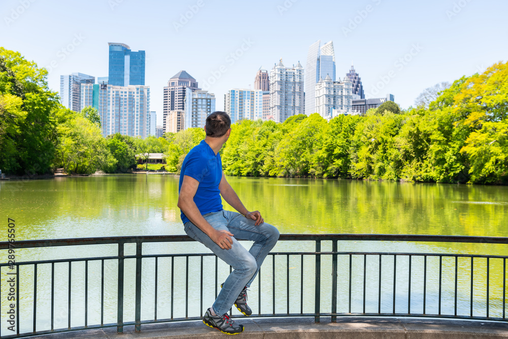 Young man sitting on railing in Piedmont Park in Atlanta, Georgia looking at scenic cityscape skyline urban view with city skyscrapers downtown by Lake Clara Meer