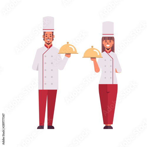 cooks couple professional chefs holding covered platters serving trays woman man restaurant workers in uniform standing together cooking food concept flat full length