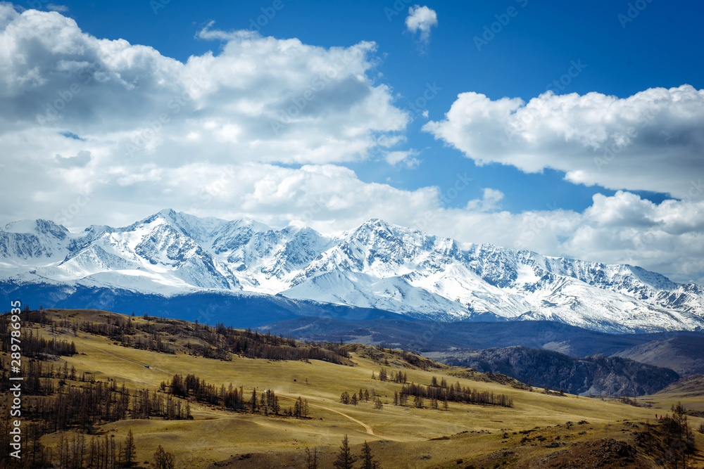 Amazing mountain landscape. Rocky mountains with snowy peaks, hills covered with grass in the Alpine scene on a bright sunny day with blue sky and clouds. View of steppe and snow-covered mountains.