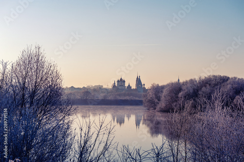 Fototapeta Naklejka Na Ścianę i Meble -  Winter landscape of temples of the city with sunset on the river Bank. The trees are covered with snow, the setting sun illuminates a beautiful mysterious landscape