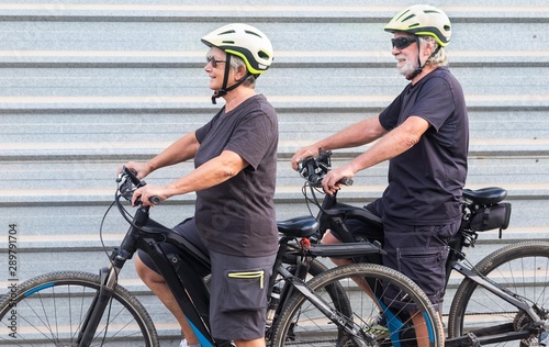 Happy senior couple doing together outdoor activity with electric bicycle. Healthy lifestyle. Yellow helmet, black clothes. Outdoor against a gray metal panel