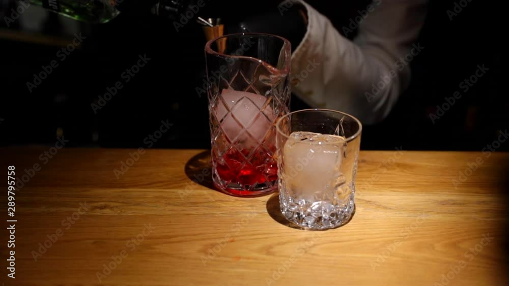 Bartender pouring alcohol into a glass using a metal measuring cup into an ice glass on a wooden bar table in light motion captured in 120fps slow motion. Bartender pouring alcohol into a glass using a metal measuring cup into an ice glass on a wooden bar table in light motion captured in 120fps slow motion.