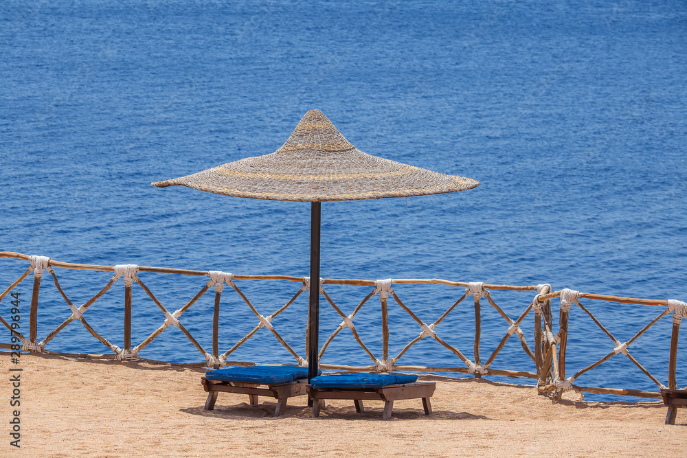 Fototapeta premium Straw umbrellas with wooden sun loungers next to the red sea on sand beach at the resort in Sharm El Sheikh, Egypt