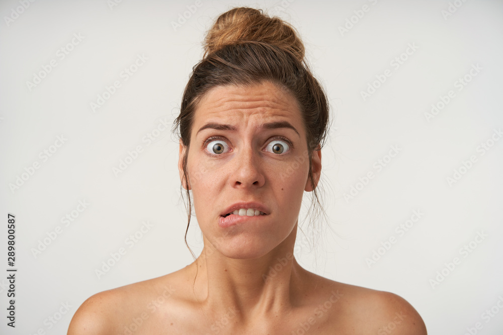 Indoor shot of young frightened woman with bun hairstyle, wrinkling forehead and biting underlip, looking to camera with wide eyes opened Indoor shot of young frightened woman with bun hairstyle, wrinkling forehead and biting underlip, looking to camera with wide eyes opened