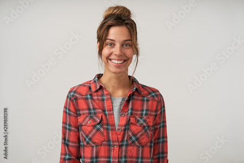 Indoor portrait of beautiful cheerful woman with bun hairstyle, looking to camera with charming smile, wearing checkered shirt, posing over white background