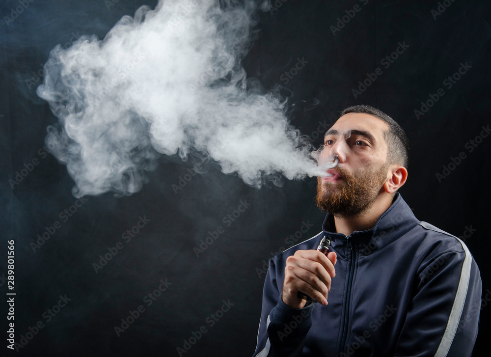 vape and smoke vape electronic e-cigarette or e-cig by a young man with smoke clouds over a black background.