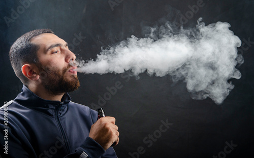 vape and smoke vape electronic e-cigarette or e-cig by a young man with smoke clouds over a black background.