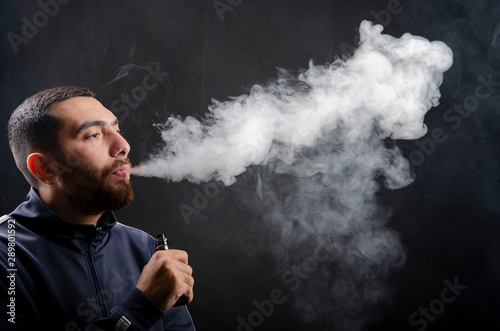 vape and smoke vape electronic e-cigarette or e-cig by a young man with smoke clouds over a black background.
