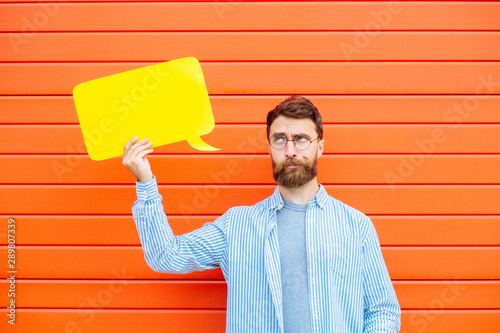 Attractive young beard man in circle glasses with fun fantasy emotion on his face holding a yellow speech bubble looking at camera and smiling, on red background outdoor portrait. photo