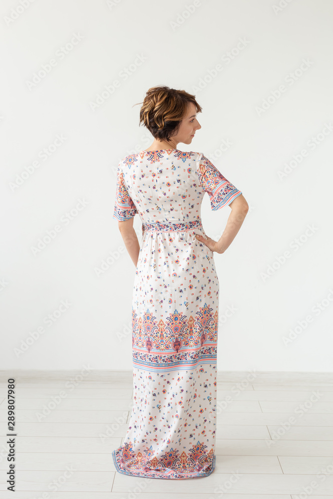 Rear view of a young slim woman in a long patterned dress standing against a white wall. The concept of unique products to order