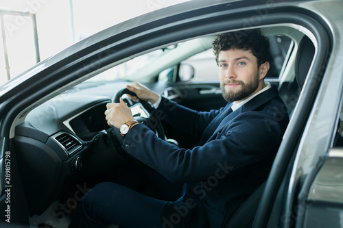 Happy car owner at the dealership. Handsome young men sitting at front seat of the car looking at camera