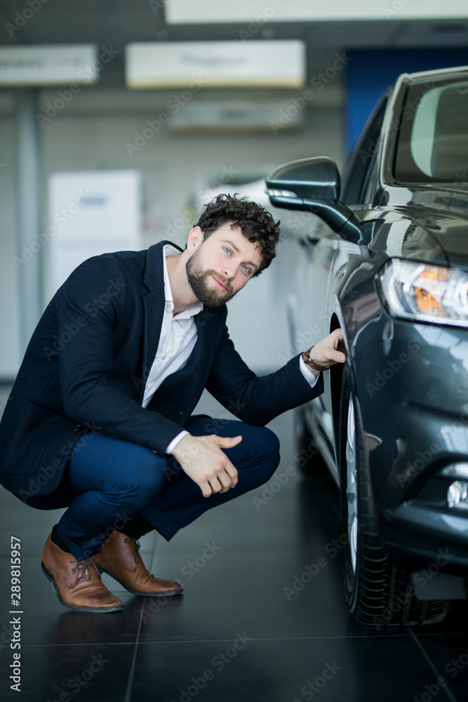 Showroom of modern car dealership. Male customer of car center holding hand on wheel of automobile. Handsome young man thinking about characteristics of new red car he want buy. Showroom of modern car dealership. Male customer of car center holding hand on wheel of automobile. Handsome young man thinking about characteristics of new red car he want buy.