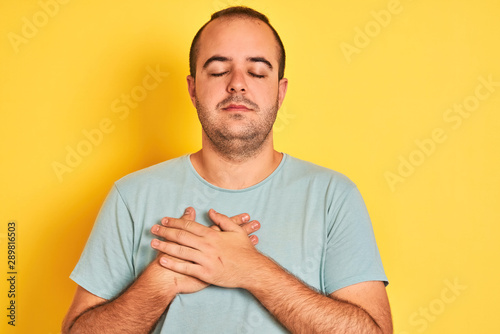 Young man wearing green casual t-shirt standing over isolated yellow background smiling with hands on chest with closed eyes and grateful gesture on face. Health concept.