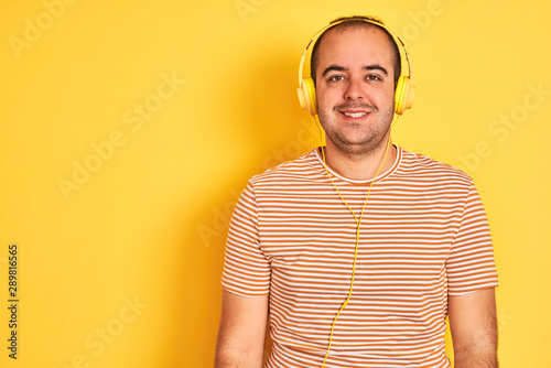 Young man listening to music using headphones standing over isolated yellow background with a happy and cool smile on face. Lucky person.