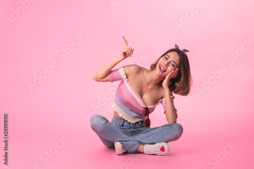 Thinking asian woman sitting on floor isolated on pink colour background.Asian female model smiling looking up.woman pointing fingers away while sitting on a floor isolated. © anon