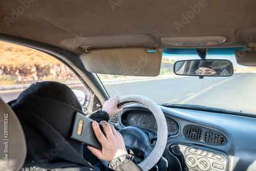 Arabic muslim woman driving a car while using her smart phone © Diya
