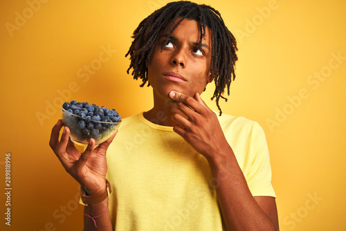 Afro american man with dreadlocks holding blueberries over isolated yellow background serious face thinking about question, very confused idea © Krakenimages.com