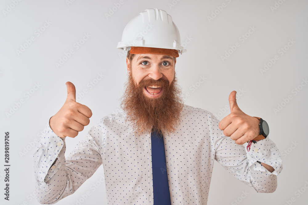 Young redhead irish architect man wearing security helmet over isolated white background success sign doing positive gesture with hand, thumbs up smiling and happy. Cheerful expression