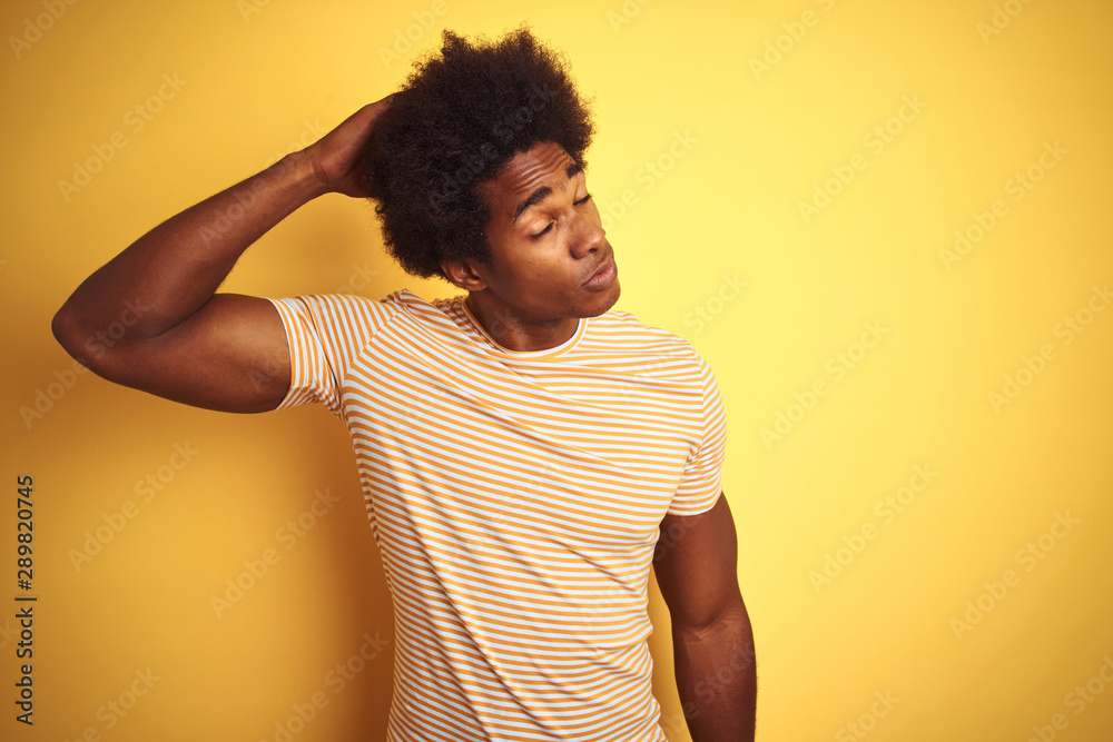 American man with afro hair wearing striped t-shirt standing over isolated yellow background confuse and wondering about question. Uncertain with doubt, thinking with hand on head. Pensive concept. American man with afro hair wearing striped t-shirt standing over isolated yellow background confuse and wondering about question. Uncertain with doubt, thinking with hand on head. Pensive concept.